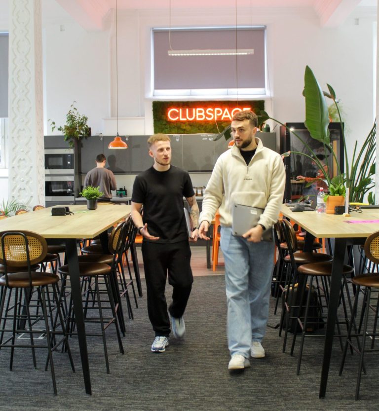 2 men walking inbetween 2 tables, 1 is holding a laptop